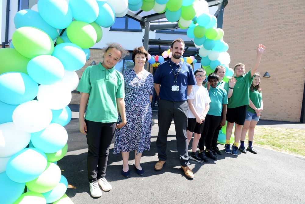 Cllr Bradwell, Headteacher and pupils standing in front of pretty balloon arch in front of new building